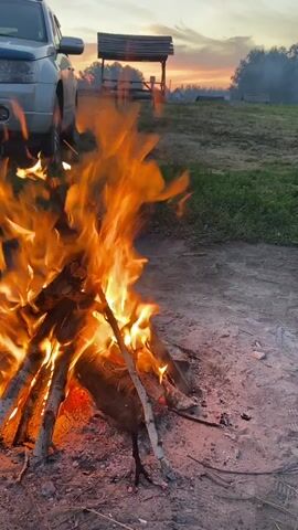 A bonfire is burning brightly, casting an orange glow on the surrounding area. A silver SUV is parked to the left of the frame, its front end visible. In the background, a wooden gazebo with a bench sits on a grassy hill under a soft, cloudy sky at dusk. The ground around the fire is covered in sand and ash. There is a brief appearance of a dog's tail and hindquarters running out from behind the SUV towards the fire.