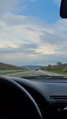 The video depicts a view from inside a car driving down a highway. The car's dashboard and steering wheel are visible in the foreground, with the road ahead stretching towards the horizon. There are other vehicles on the road, including a car in the distance. On either side of the highway, there is grassy terrain with some vegetation. In the background, there are hills or mountains visible under a sky with scattered clouds and patches of blue. There are also roadside signs and billboards. The overall scene suggests a daytime drive on a highway.