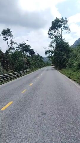 This is a vertical, first-person perspective video of a paved road winding through a lush, green landscape. The road has yellow dashed lines marking the lanes. On either side of the road are dense trees and vegetation. In the distance, a yellow vehicle is visible on the road, moving away from the camera. The sky above is partly cloudy with a mix of blue sky and white clouds. The overall impression is that of a scenic drive through a rural or natural area.