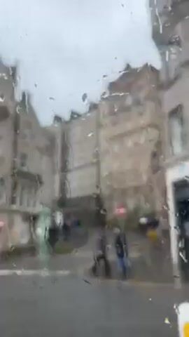 The video shows a street scene in Edinburgh on a rainy day, as viewed through a rain-streaked window. The buildings are made of gray stone and appear old, with multiple windows and architectural details. A black taxi drives past. A person walks with an umbrella.