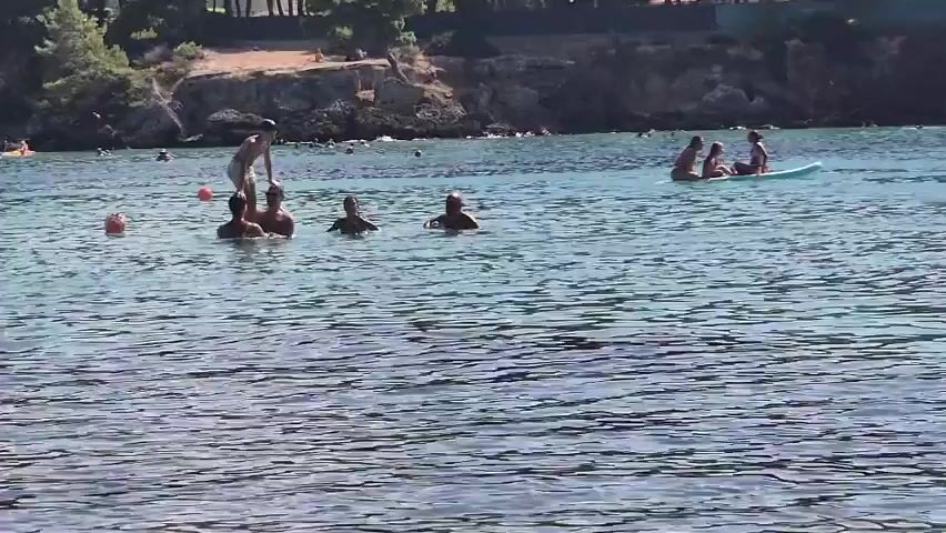 A group of people are in the ocean. In the foreground, the water is clear and rippling. In the midground, several individuals are swimming and playing. One person is doing a flip in the air, creating a large splash. Another person is sitting on a paddleboard with two other people. In the background, the shoreline is visible with trees and a hill. Other people can be seen on the beach and in the water further away. The sky is not visible.