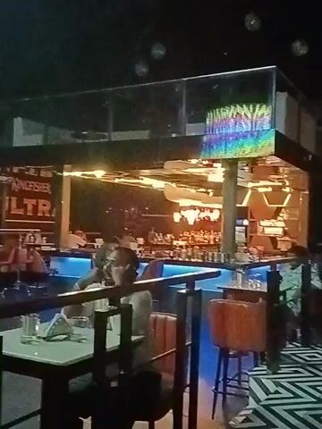 The video shows the interior of a restaurant or bar, with a multi-tiered layout and a blue-lit bar area. People are seated at tables. A large, decorative chandelier hangs from the ceiling, displaying a range of colors. There is a sign for "Kingfisher Ultra" visible on one of the walls. A few people walk around in the background. The overall lighting is dim, contributing to a relaxed or nightlife atmosphere.