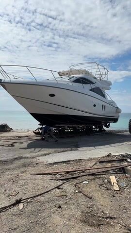 The video shows a large white yacht resting on a metal platform near a beach. A man in work clothes is moving wooden planks near the base of the yacht. Behind the yacht, a red tractor with oversized wheels is parked. The background includes a view of the ocean under a blue sky with scattered clouds. Debris, including wood and metal scraps, is scattered on the ground in the foreground. The scene appears to be a boatyard or a similar facility for yacht maintenance and storage.