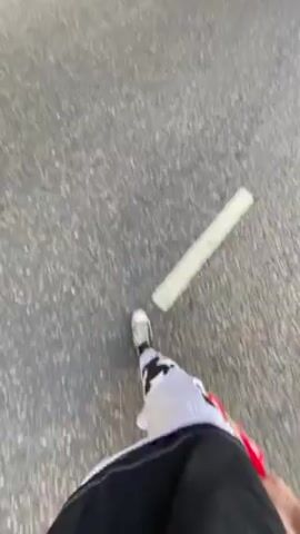 The video shows a person walking down a street with white dividing lines and parked cars. The camera angle is low, showing the person's feet and lower legs wearing white pants and different colored sneakers. As the person walks, the camera pans up to reveal a street with trees, buildings, and parked cars. There are multiple people in dark clothing standing near a dark SUV with its rear door open. The people seem to be uniformed officers. The general environment appears to be an urban setting on a sunny day.