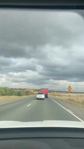 This is a first-person perspective video taken from inside a vehicle, looking forward through the windshield. The vehicle is traveling on a multi-lane paved highway. In front of the vehicle, a white passenger car is visible in the right lane, followed by a red dump truck in the same lane. The road is bordered by dry grass and some sparse vegetation on both sides. In the distance, there are utility poles and what appear to be wind turbines. The sky is heavily overcast with dark, dramatic clouds. Several yellow triangular road signs are visible along the right side of the highway, including one with an exclamation mark and another indicating a speed limit of 70. The dashboard of the vehicle is visible at the bottom of the frame.