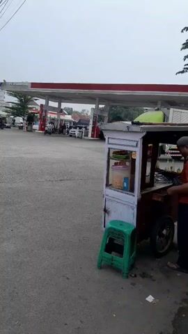 A light green truck is turning right at a gas station with red and white awnings. A small, white food cart with a green stool in front of it is in the foreground. The sky is overcast.