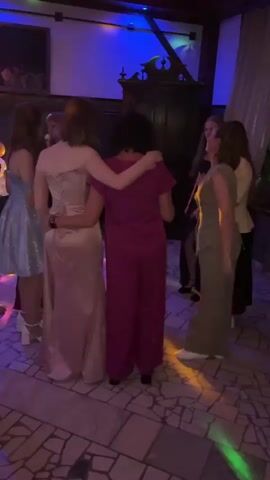 In Zavod Mikhailovskii, Russia, a group of young women are captured in a moment of celebration, likely a dance or party. The scene is set indoors, within a dimly lit room adorned with decorative wooden furniture and a large, dark wooden cabinet. The ambiance is festive, illuminated by vibrant blue and multi-colored disco lights that cast dynamic patterns on the ceiling and floor. The floor itself is tiled with a mosaic pattern in shades of grey and white.

The subjects are a group of approximately six to seven young women, dressed in formal attire. One woman in the foreground, facing away from the camera, is wearing a striking fuchsia jumpsuit. She has her arms around two other women, one in a shimmering champagne-colored gown with a corset bodice, and another in a light blue, short, sparkly dress. To the right, another woman is visible in a muted green outfit, and further back, others are partially obscured. The women appear to be engaged in dancing or interacting closely, with smiles and embraces suggesting a joyful and connected atmosphere.

The lighting and overall mood suggest the event is taking place in the evening or at night. The presence of disco lights strongly implies a celebratory occasion, possibly a prom, a birthday party, or a formal gathering. There is no visible text in the media. The overall impression is one of youthful exuberance and shared happiness within a lively social setting.