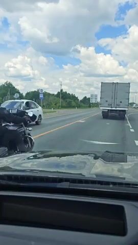 A rear-view video captures a motorcycle traveling on a paved road under a partly cloudy sky. The motorcycle is ridden by a person wearing a helmet and riding gear, with luggage secured to the rear. The motorcycle is in the right lane of the road, and a large truck is in the left lane ahead of it. Another truck is visible in the distance. The video appears to be recorded from the perspective of a vehicle following the motorcycle.