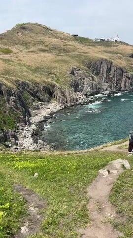 A person stands on a grassy cliff overlooking a rocky coastline with clear turquoise water. In the distance, a white lighthouse is visible atop a hill. The camera pans across the landscape, showing the rugged cliffs and the vast expanse of the ocean. The sky is partly cloudy.