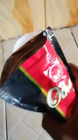 A person is pouring a bag of coffee grounds into a white plastic bucket. The bucket sits on a tiled surface. The bag has a red and black design.