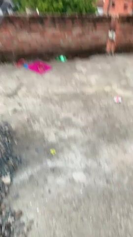 The video shows an outdoor scene from a rooftop overlooking a city. In the foreground, a brick wall runs across the screen, with a gray, concrete rooftop surface below. Scattered on the rooftop are a pink kite, two pink sandals, and a green object that appears to be a bottle. A thin string extends upwards from the kite, presumably connected to a kite being flown in the sky. In the background, a collection of buildings, trees, and a cloudy sky are visible. The camera pans slightly, capturing the scene and the distant landscape.