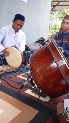 Two men are playing drums. One man on the left wears a white shirt and a blue cap and plays a small drum with his hands. The other man on the right wears a blue patterned shirt and a brown cap and plays a large, reddish-brown drum. The drums are set up with microphones. They are on a patterned rug, and a wall is visible in the background.