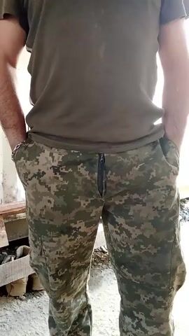 A man wearing a green t-shirt and camouflage pants stands with his hands in his pockets. His arms are slightly muscular. The camera angle is from the waist up. He is standing outside, as indicated by the light and the presence of wooden planks and a container wall. Someone walks into the frame, partially obscuring the view. The man in camouflage pants is the primary focus.