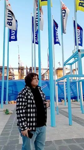 A person with short brown hair wearing a black-and-white plaid jacket, a black shirt, and blue jeans stands on a grey patterned pavement and looks up at numerous flags attached to blue poles. The flags are predominantly blue, white, and yellow and feature the word "BUILD". In the background, there's a building structure covered with blue tarp. The sky above is clear and blue. The person holds a mobile phone in their left hand.