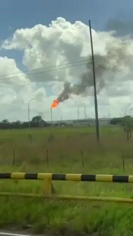The video shows an industrial area with an active gas flare. The flare is emitting a large flame and black smoke, which billows into the partly cloudy sky. In the foreground, a green field leads to the industrial facility, which is set further back. Several trees and power lines are visible between the camera and the flare. The camera seems to be moving, likely from inside a vehicle, as indicated by a glimpse of a yellow guard rail at the beginning of the video. The overall environment appears to be rural or suburban, given the extensive grassy areas.
