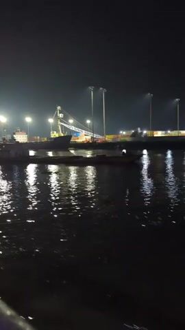 The video shows a nighttime view of a port with several boats and cargo ships docked. Large cranes are visible, suggesting ongoing cargo operations. The water reflects the bright lights from the port, creating a shimmering effect. The overall atmosphere is one of industrial activity continuing after dark.