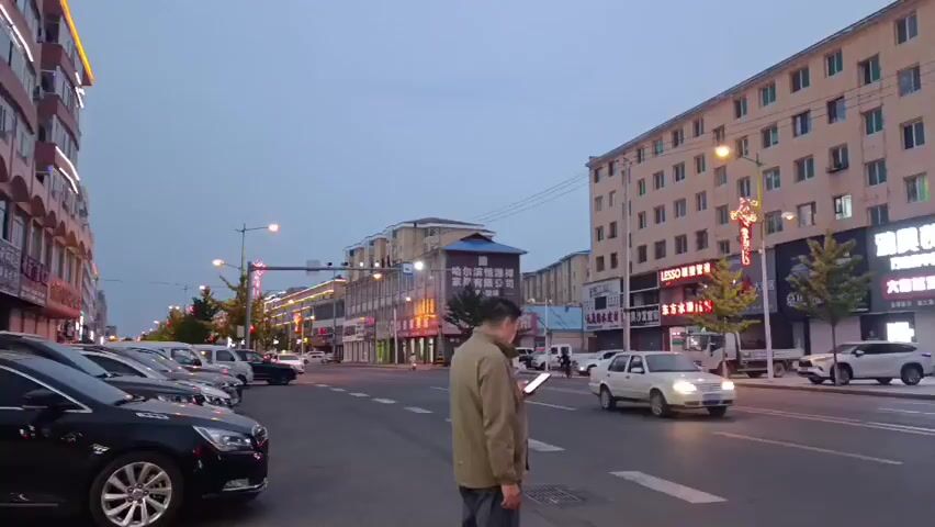 The video shows a street scene at dusk. On the right side of the street, a multi-story building with many windows lines the sidewalk. Numerous shops with illuminated signs are located on the ground floor of the building. Various cars are parked along the curb in front of the shops. A few trees are planted between the sidewalk and the street. A man is standing on the sidewalk in the foreground, looking at his phone. Cars are driving on the street, and traffic lights are visible in the distance. The sky is overcast, indicating either early morning or late evening.