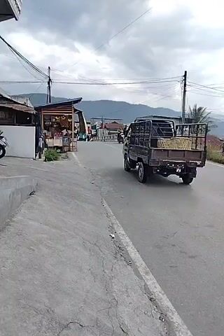 The video captures a street scene with a truck driving away from the camera on the right side of the road. A motorcycle passes the truck on the left, with a rider wearing a red shirt. The surroundings include buildings with awnings and displays of goods, power lines, a cloudy sky, and mountains in the distance. A concrete curb and a cracked asphalt road are also visible.