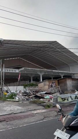 A person wearing a green shirt, black helmet, and riding a black motorcycle drives past the camera on the left. To the right of the frame, in the foreground, is a large pile of rubble, including pieces of wood, metal, and other debris. Behind the pile is a large, white building with an open, covered area supported by pillars. An Indonesian flag flies from a pole in front of the building, partially obscured by the rubble. The sky is overcast.