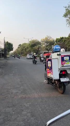 A motorcycle with a large white box attached to the back is traveling down a paved road. The box has colorful graphics on its sides and a person is riding the motorcycle, wearing a blue helmet. There are other motorcycles and a car on the road, as well as trees and buildings along the side. The sky is clear and the sun appears to be low in the sky.
