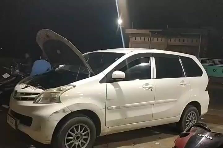 The video shows a white car with its hood open at night. A person in a blue shirt is partially visible working on the car's engine. The car is parked outdoors, possibly in a garage or workshop, with a motorcycle in the foreground. Lights are visible in the background, illuminating the scene. The overall impression is that the car is undergoing some form of maintenance or repair.