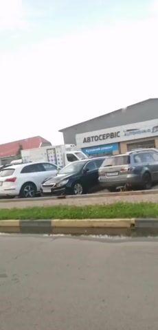 The video shows a parking lot in front of a building labeled "AUTOSERVICE" in Ukrainian. Several cars are parked in the lot, including a white SUV, a dark sedan, and other vehicles. The sky is visible with clouds. The camera pans slightly to the right and appears to be recording from the perspective of someone standing near a road.