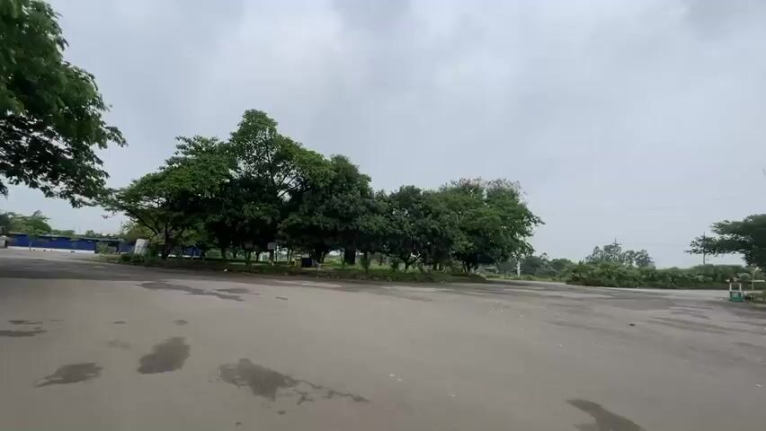The camera slowly pans across a paved circular road in what appears to be a parking lot or large driveway area. The sky is overcast. In the center of the circle are several mature trees with dense green foliage.  The pavement has cracks and patched areas. In the distance are a few small buildings and utility poles.  There are no people visible in the scene. The overall impression is of a quiet, somewhat neglected area.