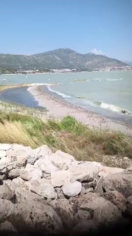 The video shows a scenic view of a body of water, likely a bay or lagoon, with a narrow strip of land extending into it. In the foreground, there is a pile of rough stones, possibly part of a wall or natural rock formation. Beyond the stones, dry grass and vegetation are visible. The strip of land is a beach, with sand and scattered debris, separating the water. The water is a light blue-green and appears somewhat choppy, indicating wind or waves. In the distance, a range of hills or mountains rises, with buildings scattered on their slopes, suggesting a coastal town or village. The sky is clear and blue, indicating a sunny day. There are no people clearly visible in the frame, but there are a few small, indistinct objects in the water that could be boats or buoys. The overall impression is a natural, somewhat wild coastal landscape.