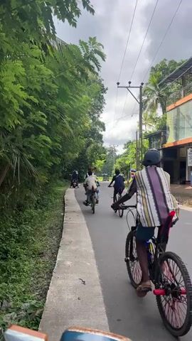 The video shows a group of young men riding bicycles down a road lined with trees and small buildings. The camera appears to be mounted on the handlebars of another vehicle, giving a point-of-view perspective of following the group. The cyclists are riding single file along the right side of the road. A three-wheeled vehicle drives in the opposite direction, passing the cyclists. The sky is overcast, suggesting a cloudy day. The environment appears to be a small town or village, with the buildings and foliage indicating a tropical or subtropical location.