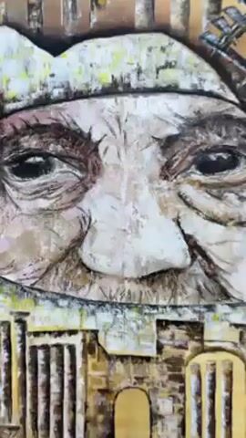The video features a large, textured painting displayed on a wooden easel. The painting is primarily rendered in earthy tones of yellow, brown, white, and black, with a heavily impastoed or textured surface.

The central and dominant feature of the painting is a large, expressive face, possibly of an older person, with deep-set eyes, prominent wrinkles, and a textured nose. The face appears to be wearing a light-colored head covering, possibly a turban or cap, with visible brushstrokes and textures.

Integrating with and surrounding the face are architectural elements. To the left, there are vertical structures resembling ancient columns or pillars. To the right, a large circular structure with spokes, characteristic of a traditional water wheel (noria), is depicted. Below the face, the painting shows more architectural details, including stylized arches, rectangular building blocks, and what appears to be fragmented structures or ruins, all rendered with similar rough textures and a limited color palette.

As the video pans out, the full scope of the painting on the easel is revealed. In the background, behind the easel, another large artwork is visible on a wall. This background piece is more colorful, featuring a vibrant blue upper section and a green lower section that resembles a stylized map, possibly of a geographical region or country. On the blue section, there are several white shapes, possibly birds or leaves, some with what appears to be black Arabic script written on them.

The floor beneath the easel is tiled with a pattern of light and dark brown or beige square tiles. The overall setting suggests an indoor space, possibly an art gallery, studio, or exhibition area. The video slowly zooms out from a close-up of the painting's face to a wider shot, showcasing the entire artwork and its immediate surroundings.