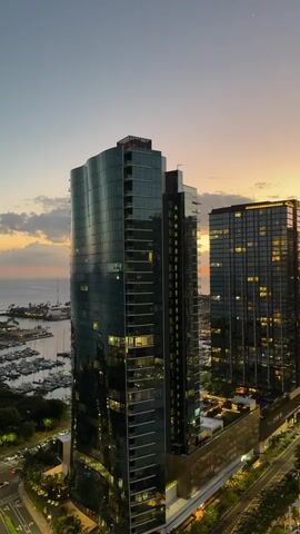 This video shows an aerial view of two modern skyscrapers with glass facades at dusk. The buildings are lit up from within, creating a warm glow. In the foreground, to the left, is a body of water with a marina filled with sailboats. Behind the skyscrapers, the sky is a gradient of soft orange, yellow, and blue, with a few wispy clouds. The overall scene is one of urban development blending with a coastal landscape. There are no people visible in the video.