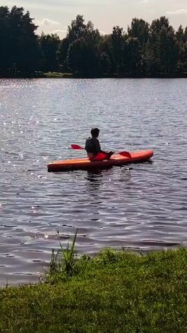 A person is kayaking on a lake. The kayak is orange and the person is wearing a life jacket and red shorts. The person is paddling with a red paddle. The lake is calm and the sun is shining, creating a shimmering effect on the water. There are trees in the background and green grass in the foreground. The video is shot from a distance.