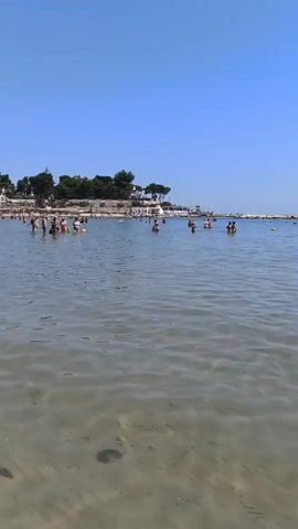 A bright, clear day at a beach. The camera is positioned low in the water, looking out towards a crowded shoreline. The water is shallow and clear, with the sandy bottom visible. In the foreground, there are two red paddleboards floating, one with a woman sitting on it. Further out, numerous people are swimming and wading in the water. In the background, trees line the shore, and a stone breakwater extends into the sea. The sky is a vast, cloudless blue.