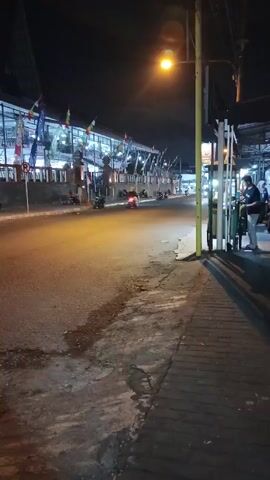 The video captures a nighttime street scene. A lamppost with an illuminated bulb stands to the right of the frame, casting a warm glow on the asphalt road. The road is wet in patches, reflecting the light. On the left side of the road, a long building with a covered walkway is visible, decorated with numerous small flags and banners. Several motorcycles are present, with two moving vehicles on the road and a few parked. On the right side of the road, a sidewalk is paved with bricks. Two people are standing near a street vendor's cart or stall. One person, wearing a dark shirt and light pants, is standing, looking down at something, possibly a mobile phone. Another person, wearing a dark t-shirt and jeans, is walking away from the camera. The overall atmosphere is dimly lit and appears to be in a commercial or residential area.