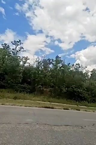 The video shows a roadside view with a cloudy blue sky and trees lining the road. The camera appears to be moving, likely from within a vehicle. The road itself is visible in the foreground and shows some cracks and wear. The trees are dense and green, forming a natural barrier along the side of the road. The clouds are white and fluffy, indicating fair weather. There are no people or other vehicles clearly visible in the frame.