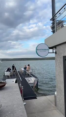 The video captures a serene view of a gastropub named "ALTA MARE" situated by a lake. The gastropub features a floating platform extending into the water, where patrons are seen dining and enjoying the view. Some are seated at tables, being served by a waiter, while others are standing near the railing, taking in the scenery. The sky is overcast with a mix of grey clouds and patches of blue. In the distance, a sailboat glides across the lake. On the pier leading to the floating platform, there's a fire pit and a grey mat flanked by red ropes, indicating a designated walkway. The overall ambiance suggests a tranquil and scenic dining experience.