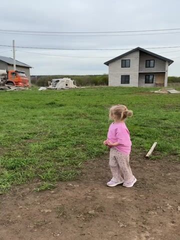 A little girl is standing in a yard, looking at a concrete mixer truck. She is wearing a pink shirt and patterned pants. The yard is grassy with patches of dirt. There are two houses in the background. The sky is overcast. The truck is orange and white and has "Beton" written on the side. The girl turns and smiles.