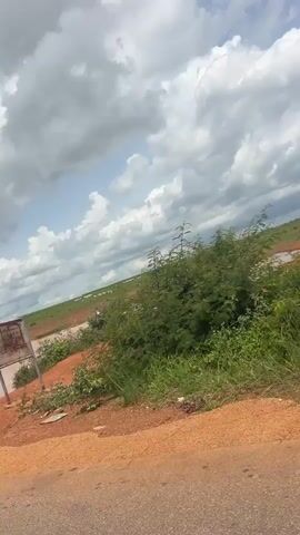A video shows a landscape with a large body of muddy water. The water is surrounded by green grass and red earth. In the distance, a few white objects, possibly livestock, are visible in a field. The sky is partly cloudy. The camera appears to be moving, possibly from a vehicle, as the foreground shows a dirt road. The overall impression is of a rural or undeveloped area.
