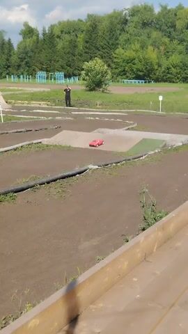The video displays an outdoor race track constructed of dirt with ramps. In the background, there are trees and playground equipment. Two individuals are present on the track. One person, wearing a black shirt and dark pants, stands near a ramp, seemingly observing the race. Another person, in a blue shirt and dark shorts, is positioned further away, also appearing to be a spectator. Several remote-controlled cars are racing on the track. A red car is seen in the initial frames, followed by a green car. The video captures the cars navigating the track and its obstacles.