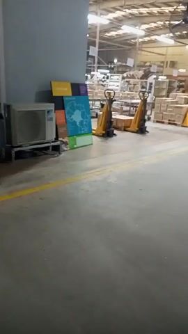 The video shows an interior view of a warehouse or factory floor. The floor is concrete with yellow safety lines. Several yellow manual pallet jacks are scattered throughout the space, some empty and others loaded with pallets of cardboard boxes. There are also shelves filled with various items and a few metal carts. In the foreground, there is a standing sign with sections of different colors (yellow, purple, orange, brown, blue, and green). The blue section features a map of Africa. A Daikin air conditioning unit sits next to the sign. The overall lighting is bright, and the warehouse appears to be well-organized. There are no people visible in the shot.