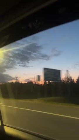 The video shows a view from inside a car driving on a highway at dusk. A lamp post illuminates the road. The sky is a mix of blue and orange hues. A line of trees is silhouetted against the sky.