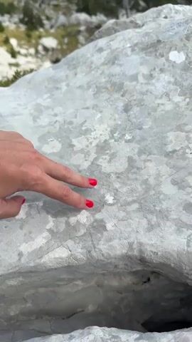 The video begins with a close-up of a person's hand with bright red nail polish touching a large, textured grey rock. The hand is positioned on the left side of the frame, and the rock fills most of the foreground. The background is out of focus but appears to be a natural, rocky environment with some greenery. The camera then pans up and zooms out to reveal a breathtaking panoramic view of a mountainous landscape. Lush green forests cover the slopes of numerous mountains that recede into the distance, fading into a hazy blue. The sky is partly cloudy with white, fluffy clouds against a bright blue sky. The sun appears to be shining, casting light and shadows across the rugged terrain. The scene conveys a sense of vastness and natural beauty.