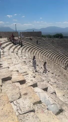 This media file captures a wide-angle view of an ancient amphitheater in Pamukkale, Turkey. The scene is bathed in bright sunlight, indicating a clear, warm day, likely mid-morning or early afternoon given the shadows. The sky is a vibrant blue with scattered white clouds, and in the background, distant, hazy mountains form a picturesque backdrop.

The amphitheater itself is the central focus, constructed from weathered, light-colored stone. Its tiered seating curves in a vast semi-circle, with rows of steps ascending towards the upper levels. The architecture suggests a historical site, likely a well-preserved ancient Roman or Greek theater. Wooden barriers are visible along some of the pathways and edges of the seating area, indicating that it is a tourist attraction.

Several people are scattered throughout the amphitheater. In the foreground, two women in casual clothing are walking down the stone steps. Further back, groups of tourists are seen seated or walking on the various levels of the seating area. Some individuals are also visible near the stage area, which is partially visible at the bottom right of the frame. The people appear to be casually exploring and enjoying the historical site.

The overall atmosphere is one of serene exploration and appreciation for ancient history. The bright sunlight and clear sky contribute to a positive and inviting mood. The surrounding landscape, with its dry, earthy tones and distant greenery, adds to the sense of being in a historical and scenic location. No visible text is present in the media file.