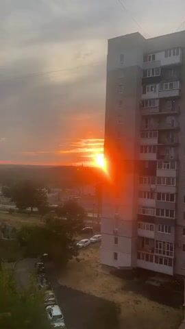This is a vertical video clip of a sunrise over Kropyvnytskyi, Ukraine. The sun is a bright, intense orb in the sky, casting a strong orange and yellow glow. Rays of light emanate from the sun, creating a lens flare effect that streaks across the right side of the frame, illuminating the side of a tall, multi-story apartment building. The building, with its repeating pattern of windows and balconies, occupies the right portion of the image.

In the background, a landscape stretches out, appearing somewhat hazy due to the sunrise. There are some trees, buildings, and what appear to be industrial structures or chimneys visible in the distance. The sky is a mix of warm hues of orange and yellow from the sunrise, with some wispy clouds present. Below the apartment building, a road and a small parking area are visible, with several cars parked. A patch of dry, brown grass is also noticeable in the foreground.

The time of day is clearly sunrise, indicated by the position and intensity of the sun and the warm colors in the sky. The weather appears to be clear, though the haziness in the distance might suggest some atmospheric moisture. The overall mood evoked is one of the beginning of a new day, with the vibrant sunrise creating a visually striking and somewhat dramatic scene. There is no visible text in the media file.