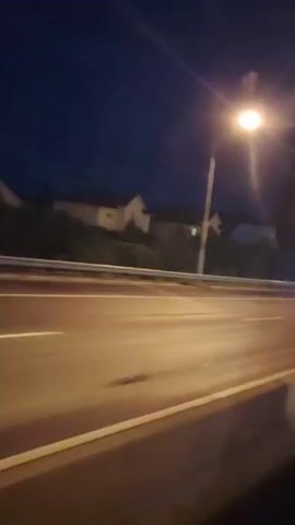 A handheld, slightly blurry video shows a nighttime view from a moving vehicle on a road. A street light illuminates the scene, casting a bright glow and creating lens flare. In the background, houses and trees are visible under a dark blue sky. The road has clear lane markings, and a guardrail runs alongside it. Several cars pass by, their headlights and taillights streaking due to the motion. One car is particularly bright with headlights on. A billboard is also visible. The camera appears to be looking forward and slightly to the right, capturing the flow of traffic.