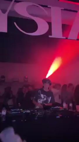 A young DJ is mixing music at a party or club. He has long dark hair pulled back into a ponytail, and he's wearing a baseball cap and a dark t-shirt with a black and white graphic on it. He’s standing behind a DJ console with lots of knobs and buttons, clearly focused on his work. The scene is filled with people, mostly blurred out in the background, who seem to be enjoying the music and atmosphere. Red and orange lights pulse, creating a vibrant, smoky feel that highlights the energy of the space. The camera is held by someone in the crowd, who’s hand can be seen in the foreground with a bracelet on the wrist. A large partially visible white sign hangs overhead.