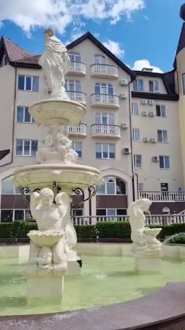 A video shows a three-tiered white fountain in front of a light-colored building with a red tiled roof and balconies. The fountain has several white statues, including a standing female figure at the top, cherubic figures in the middle, and a male figure with wings at the base. Water sprays from the fountain into a green pool. The sky is blue with scattered clouds. The fountain and building appear to be outdoors in a well-maintained area.