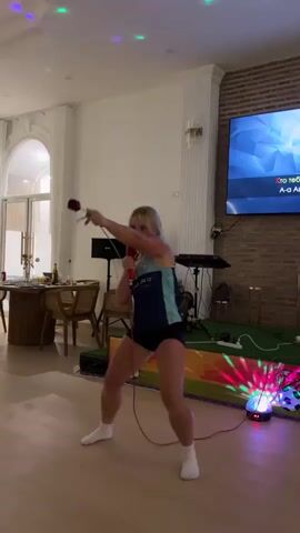 A young woman with blonde hair is holding a microphone in her right hand and a red rose in her left hand. She is wearing a dark blue athletic tank top with teal accents and black shorts, and white socks. She is standing in a room with a wooden floor and white walls. There is a large television screen in the background displaying lyrics. A disco ball is on the floor to her right, casting colorful lights on the scene. A table with chairs is visible to the left, and a brick wall is behind the television. She appears to be singing and performing.