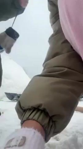 In a snowy outdoor scene, a person in a green jacket holds a small glove attached to a string, offering it to another person in a pink and green outfit. The latter, wearing white gloves, takes the glove from the first person. The ground is covered in snow, with a pile of snow in the background. Several shovels and other tools are scattered on the snow. A building is also visible in the background. The sky is overcast, suggesting a cold or wintry day.