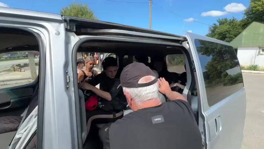 A group of people are gathered around the open sliding door of a silver van. An older man with gray hair and a black baseball cap is seen from behind, his right arm extended into the van. Inside the van, several people are seated. A woman with dark hair in a black top and black leggings is sitting in a middle seat, holding a red bag. Another woman with blonde hair, wearing a black tank top and black leggings, is leaning into the van from the doorway, holding a black bag. Two other individuals, one wearing a black hat and another with curly dark hair, are visible further inside the van. Luggage is stowed in the van. The scene appears to be outdoors on a sunny day, with a road and trees visible in the background.